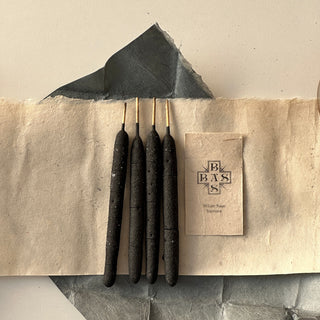 Four black hand-rolled white sage incense sticks placed on a beige paper with a cardboard packaging that has the brand 'Basbas' printed on it.
